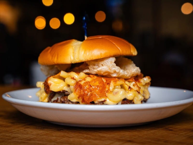 The burger features two seared beef patties topped with American cheese, creamy mac and cheese with bacon, and shredded cheese, served on a brioche bun with house-made comeback sauce and finished with crispy fried pork skins.