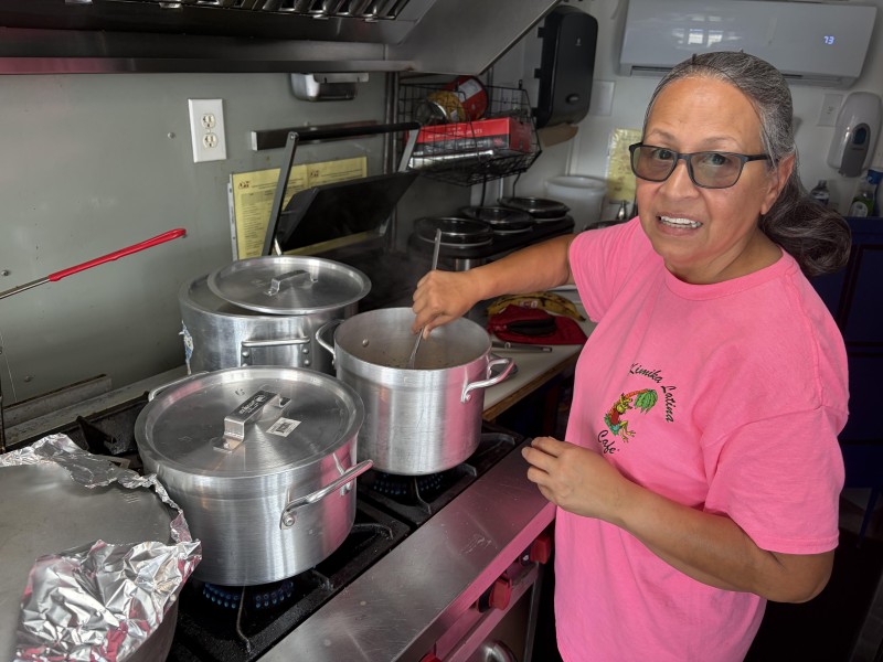 The owner is wearing a pink T-shirt with the food truck's log and is stirring a silver-colored pot inside the food truck.