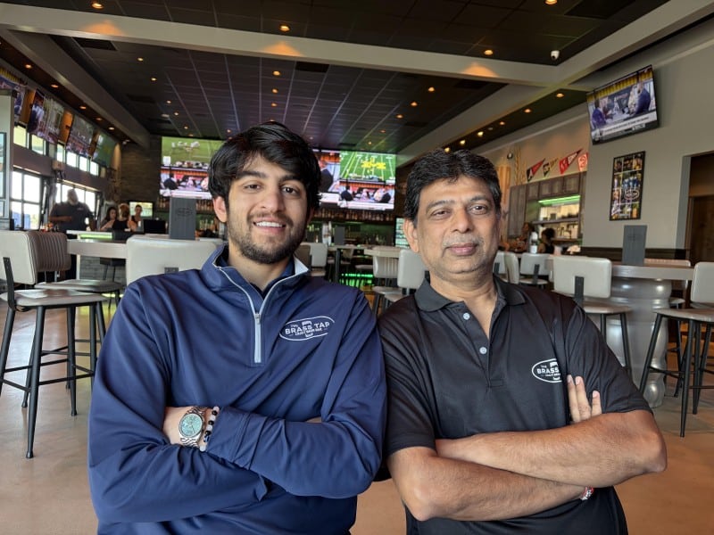 Son and father side by side inside The Brass Tap with tables, chairs, n L-shaped bar, TV screens in the background.