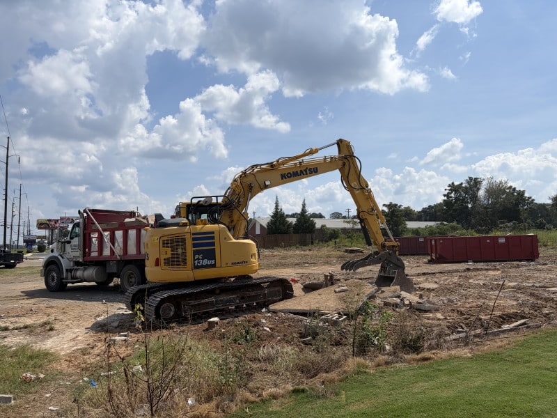 A hydraulic crawler excavator is shown removing concrete from the demolition site and place the debris in a waiting truck.