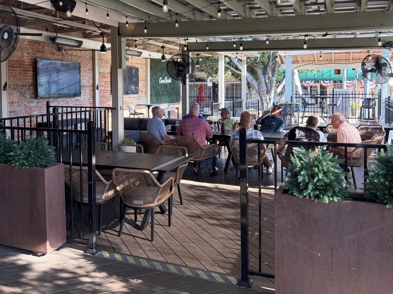 The outdoor event venue has an outdoor bar, chairs and tables for dining, a place to take a selfie and a stage for band. The group of men are seated at a long table smoking cigars and enjoying drinks and conversation.