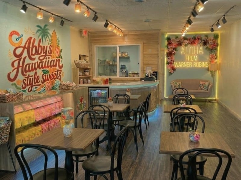 Tables for two or three with the business logo on the left wall, a counter in the left corner and selfie board with a chair underneath in the far back right. Hawaiian themed interior.