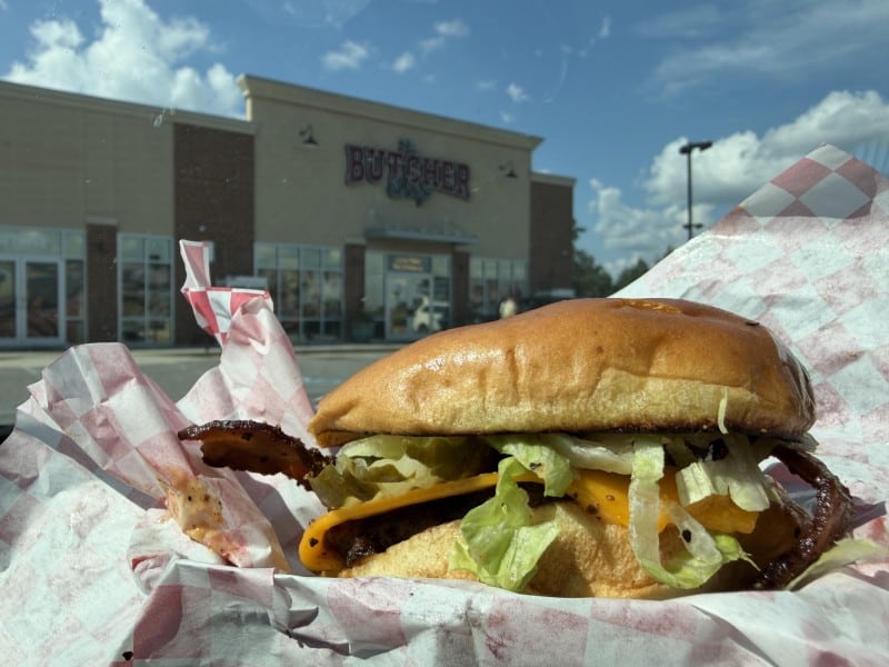The burger piled with all its trimmings of bacon, cheese, lettuce, onion is shown in the foreground with The Butcher Shop building shown in the background.