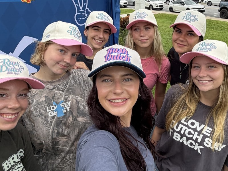Regional operator is in the center of the selfie wearing a "Rush Dutch" baseball cap surrounded by six young women wearing Dutch Bros T-shirts and "Rush Dutch" baseball caps with a blue screen in the background with the Dutch Bros Coffee logo, a peace sign and cars parked along the sidewalk on the right in the background.
