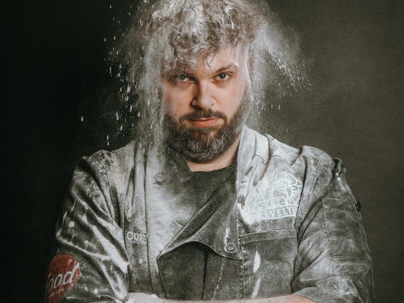 Cory is covered with flour falling overhead. He has a stern expression on his face, he's wearing a bakery uniform that's covered in flour and he has his armed crossed.