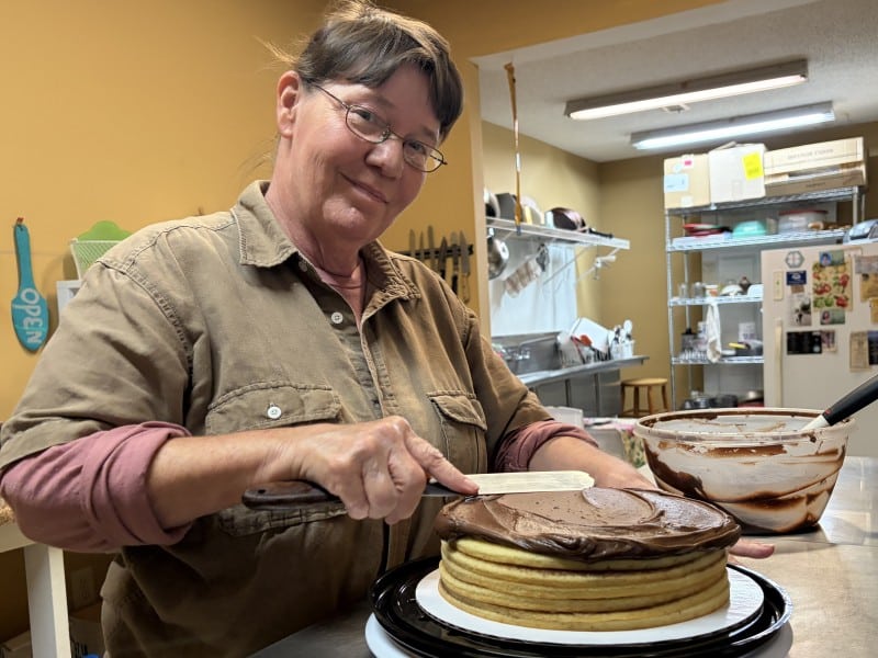 Baker ices a six layer chocolate buttermilk cake with the kitchen shown in the background.