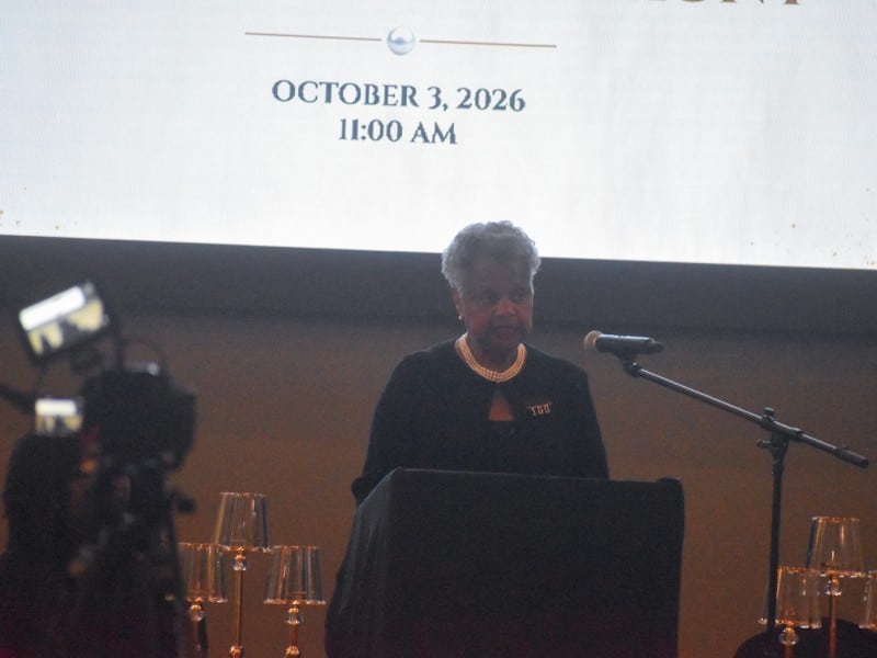 A woman in a black dress standing behind a podium. She is speaking into a microphone.