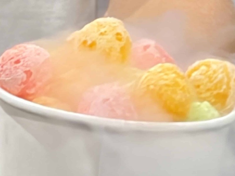 "Smoke" can be seen coming off the small orange, pink and yellow balls of the dessert. A man's black rubber gloved hand can be see holding up the dessert that's in a white paper cup.