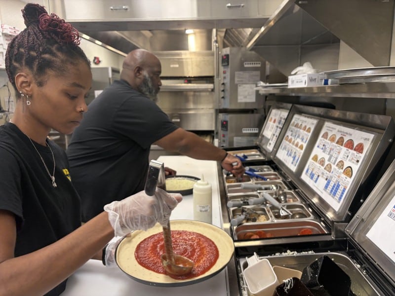 The woman and man are in front of pizza ingredients. She's spreading sauce on pizzas and he's adding the cheese and other toppings.