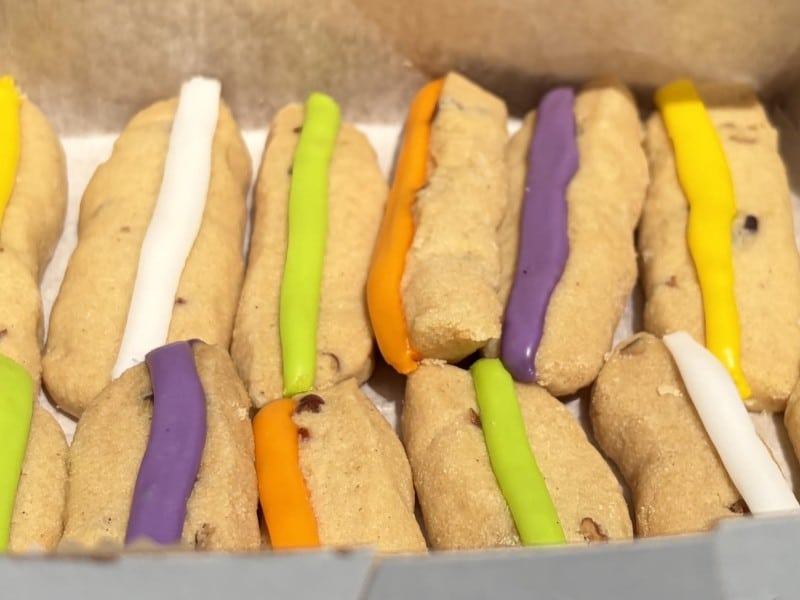 The white box is filled with the cookies that look like lady fingers with a stripe of icing in yellow, white, neon green, neon orange, yellow and purple colors. Pecans pieces baked baked in the cookie dough are evident.